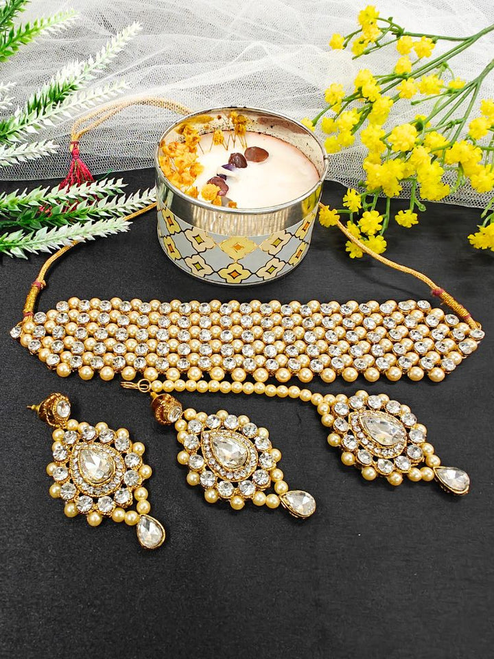 The Jayshree Champagne Necklace Set features a choker, earrings, and maang tikka adorned with pearls, American diamonds, Champagne beads—perfect for weddings. Displayed with a lit candle and yellow flowers.