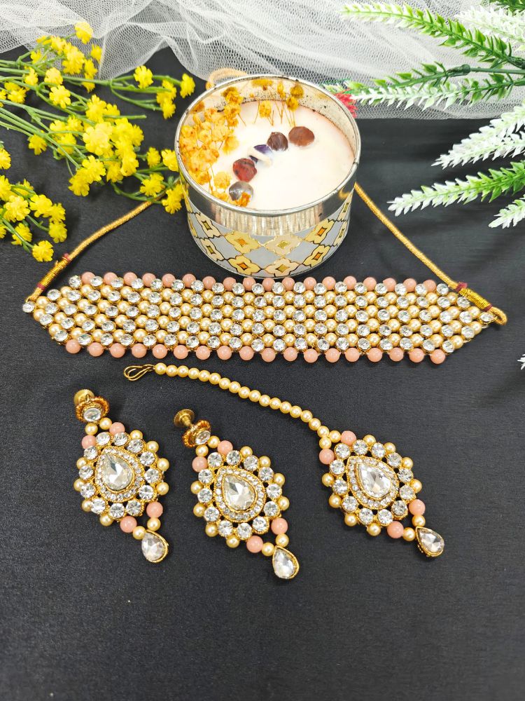 The Jayshree Peach Necklace Set features a choker, earrings, and maang tikka adorned with pearls, American diamonds, Peach & Golden beads—perfect for weddings. Displayed with a lit candle and yellow flowers.