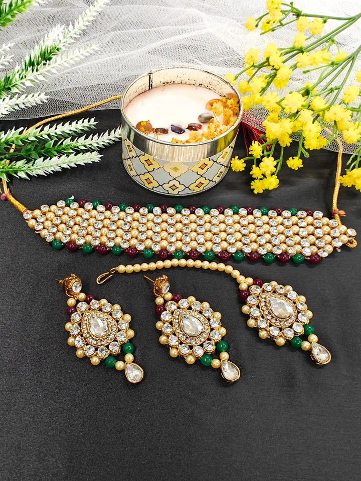 The Jayshree Red & Green Necklace Set features a choker, earrings, and maang tikka adorned with pearls, American diamonds, green and maroon beads—perfect for weddings. Displayed with a lit candle and yellow flowers.