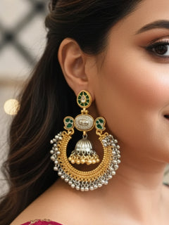A close-up of adorned with Jhanak Green Earrings, featuring ornate green stones, gold, brass, and silver details, intricate circlular patterns, small beads, pearls, and a small jhumki at the centre. Oxidized earrings, german silver earrings, navratri earrings. 