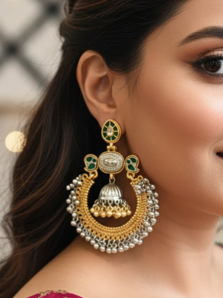 A close-up of adorned with Jhanak Green Earrings, featuring ornate green stones, gold, brass, and silver details, intricate circlular patterns, small beads, pearls, and a small jhumki at the centre. Oxidized earrings, german silver earrings, navratri earrings. 