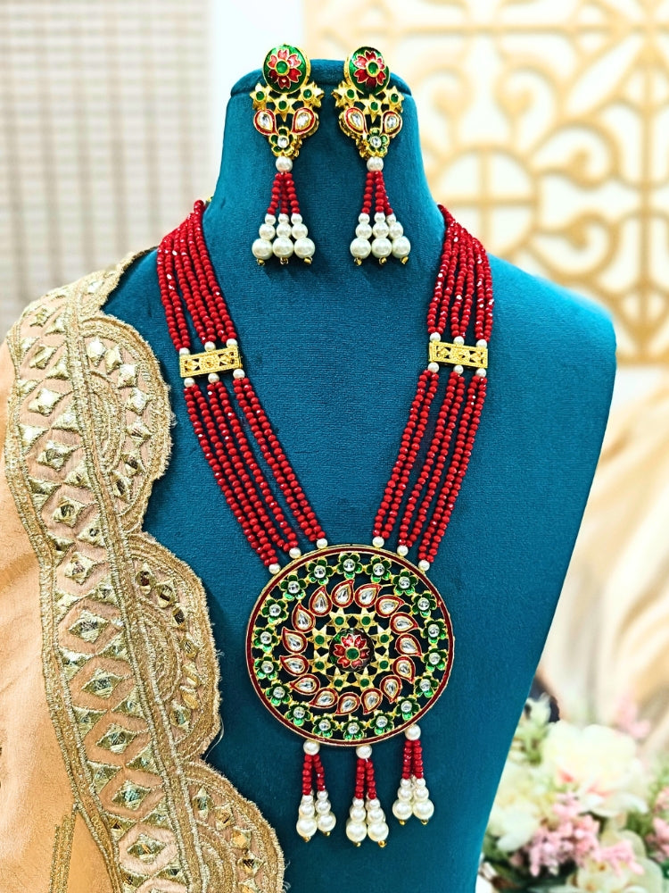 A mannequin displays the Jinisha Green Necklace Set, featuring a traditional Kundan Red necklace with a large round pendant set with red, green, and white stones, plus matching earrings accented with pearl drops. It has multi-layer strands to give a heavy look. 