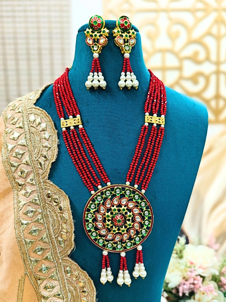 A mannequin displays the Jinisha Green Necklace Set, featuring a traditional Kundan Red necklace with a large round pendant set with red, green, and white stones, plus matching earrings accented with pearl drops. It has multi-layer strands to give a heavy look. 