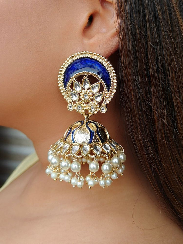 A close-up of a woman wearing the Juliette Earrings, featuring large blue kundan and meenakari earrings, with ornate pearls and gold detailing reminiscent of Chandbali style, highlights the jewelry against her ear and straight brown hair.
