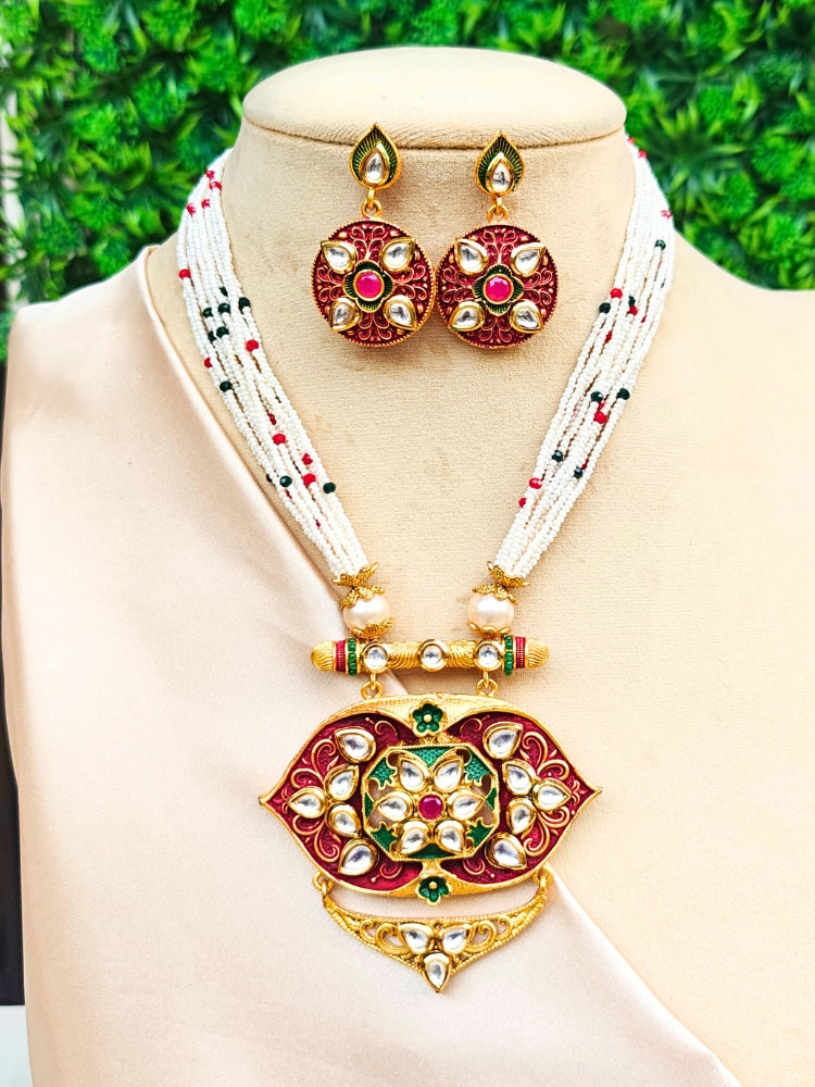 The Jwala Necklace Set features a beaded Meenakari necklace with a large gold, red, and green Kundan meenakari pendant and matching circular earrings, showcased on a mannequin against a backdrop of green leaves.
