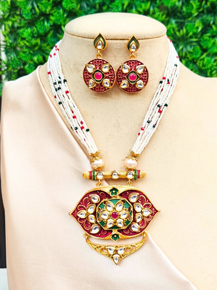 The Jwala Necklace Set features a beaded Meenakari necklace with a large gold, red, and green Kundan meenakari pendant and matching circular earrings, showcased on a mannequin against a backdrop of green leaves.