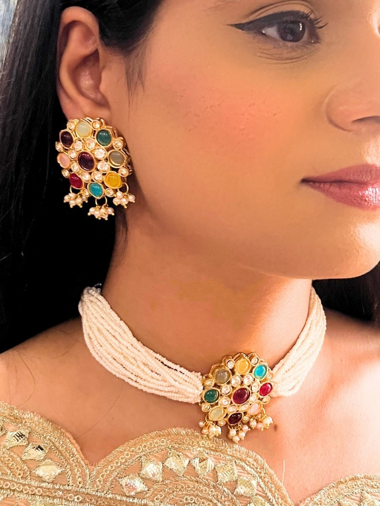 A woman wears the Kairavi Multi-color Choker Necklace Set, featuring a gold and colored stones earring with a matching pendant held by multiple pearls strands. She is dressed in a cream embroidered outfit, showing only her lower face and neck.