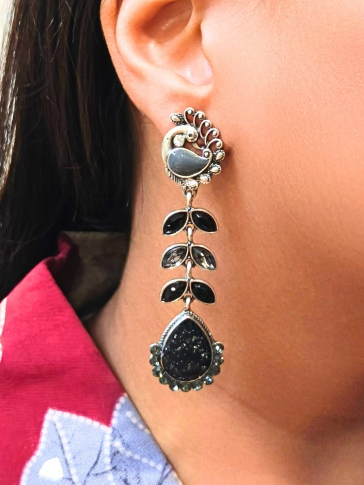 A close-up of a womans ear wearing Kajal Black Earrings—large, ornate earrings with brass and American diamond accents, leaf-shaped segments, and a teardrop pendant. She pairs them with a pink and blue patterned top—ideal Wedding Ceremony Jewelry. Oxidized earrings, german silver earrings, navratri earrings. 