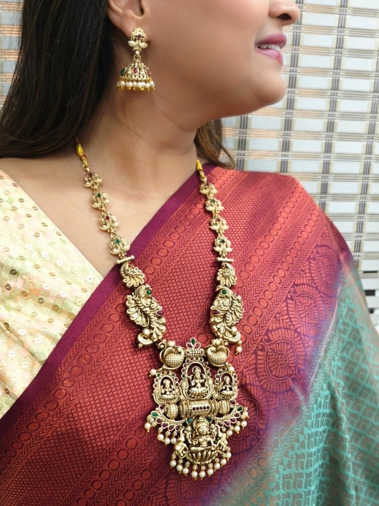 A woman wears a maroon and green sari with gold South-Indian jewelry, including the Kamakshi Temple Necklace Set featuring intricate traditional designs and deity motifs.