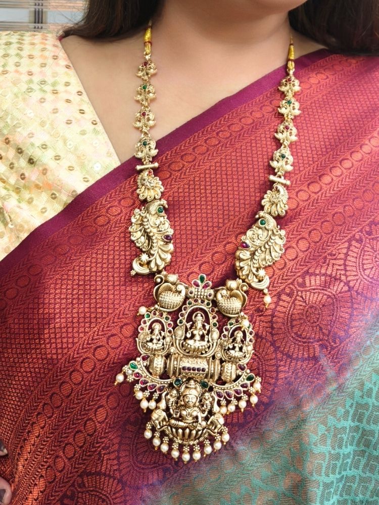 A woman wears a maroon and green sari with gold South-Indian jewelry, including the Kamakshi Temple Necklace Set featuring intricate traditional designs and deity motifs.