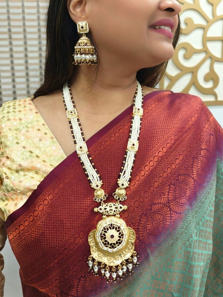 A woman in a maroon and green saree with a gold blouse wears the Kamala Necklace Set— a necklace with multiple strands of pearls, meenakari and colored stone motifs, along with a big pendant with meenakari and gold work at the bottom. Matching earrings come along with it. She smiles slightly, looking to the side, with geometric wall art behind her.