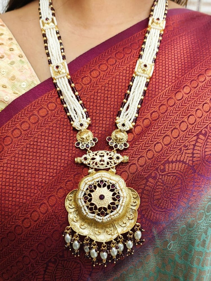 A woman in a maroon and green saree with a gold blouse wears the Kamala Necklace Set— a necklace with multiple strands of pearls, meenakari and colored stone motifs, along with a big pendant with meenakari and gold work at the bottom. Matching earrings come along with it. She smiles slightly, looking to the side, with geometric wall art behind her.