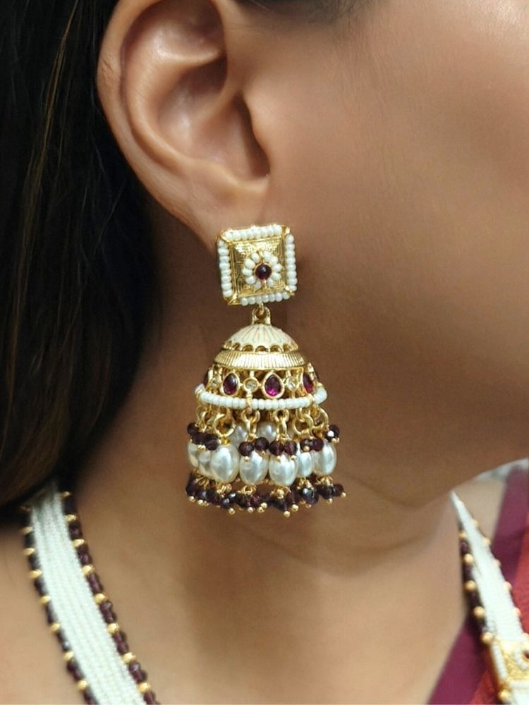 A woman in a maroon and green saree with a gold blouse wears the Kamala Necklace Set— a necklace with multiple strands of pearls, meenakari and colored stone motifs, along with a big pendant with meenakari and gold work at the bottom. Matching earrings come along with it. She smiles slightly, looking to the side, with geometric wall art behind her.