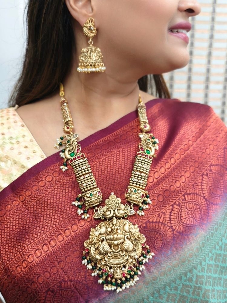 A woman wearing a rich maroon and gold saree, adorned with the Kantha Temple Necklace Set featuring intricate detailed design of  Maa Laxmi, matching wedding accessories, and elegant gold jhumka earrings.
