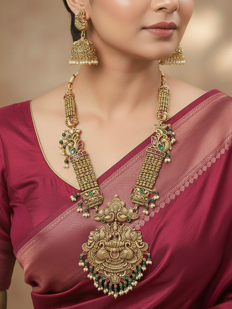 A woman wearing the Kantha Temple Necklace Set featuring a traditional gold Long necklace and matching earrings with Goddess Laxmi in the middle, along with pink and green colored stones, paired with Saree —Bride showcasing a premium temple artificial jewelry set for wedding rituals.