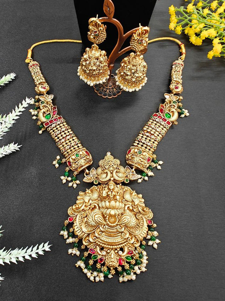 The Kantha Temple Necklace Set featuring a traditional gold Long necklace and matching earrings with Goddess Laxmi in the middle, along with pink and green colored stones, paired with Saree —Close-up of detailed temple carvings in artificial wedding jewelry.