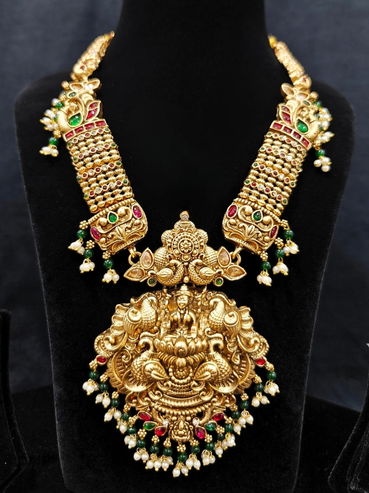 the Kantha Temple Necklace Set featuring a traditional gold Long necklace and matching earrings with Goddess Laxmi in the middle, along with pink and green colored stones, paired with Saree —Bride showcasing a premium temple artificial jewelry set for wedding rituals.