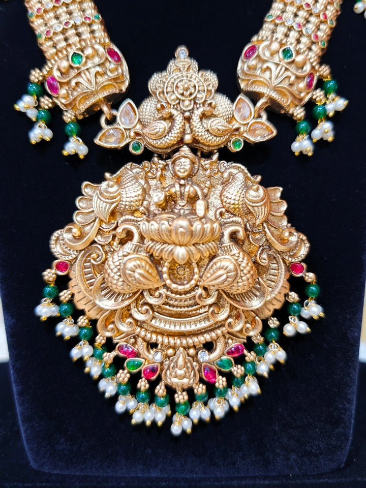 the Kantha Temple Necklace Set featuring a traditional gold Long necklace and matching earrings with Goddess Laxmi in the middle, along with pink and green colored stones, paired with Saree —Bride showcasing a premium temple artificial jewelry set for wedding rituals.