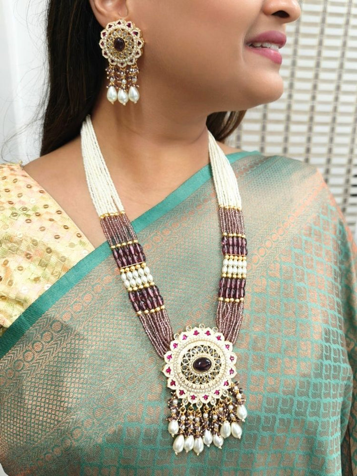 A woman wears the Kashish Necklace Set— A necklace with multiple strands of white pearls and pink crystal stones, with a big pendant made with enamel meenakari work, kundan detailing and an oval pink-purple stone in the middle, along with pearls and other crystals hanging at the bottom. Only her lower face and upper torso are shown; she’s dressed in a green and gold saree. Close-up of enamel work in Meenakari artificial wedding jewelry.