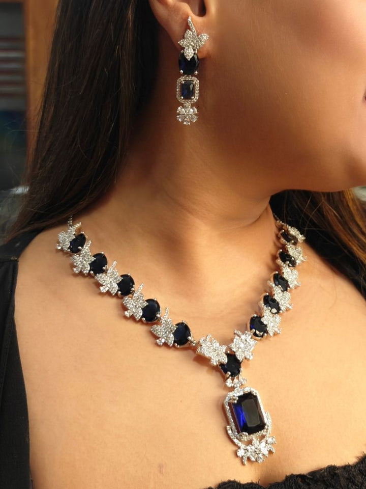 A woman wears the Peacoski Katrina Blue Necklace Set, featuring a necklace and matching dangling earrings adorned with large Blue stones and sparkling American diamonds in ornate silver settings.