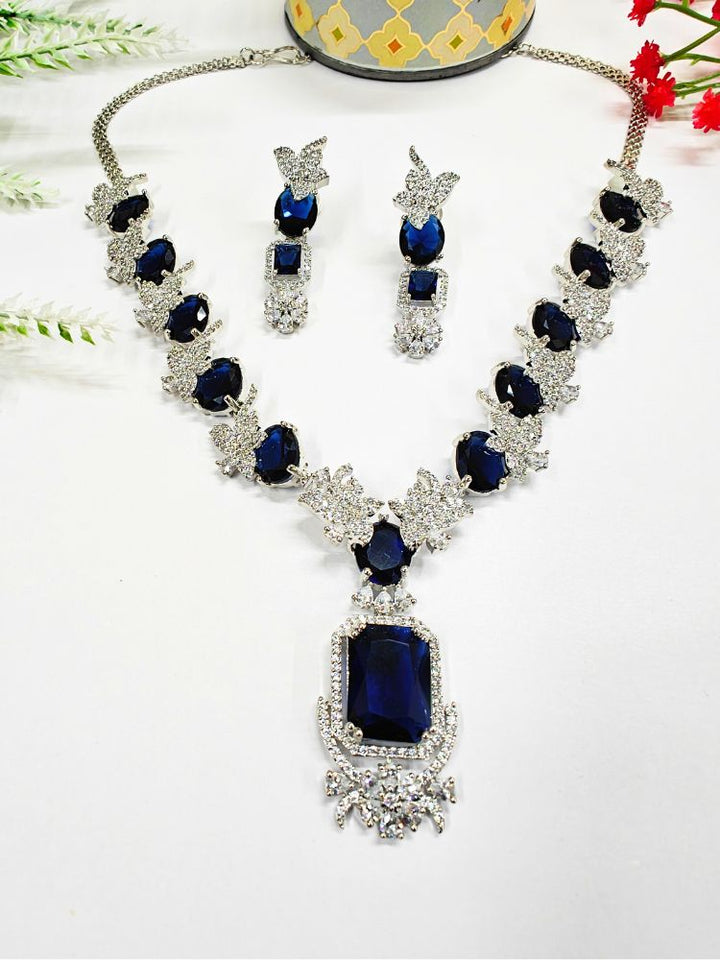 A woman wears the Peacoski Katrina Blue Necklace Set, featuring a necklace and matching dangling earrings adorned with large Blue stones and sparkling American diamonds in ornate silver settings.