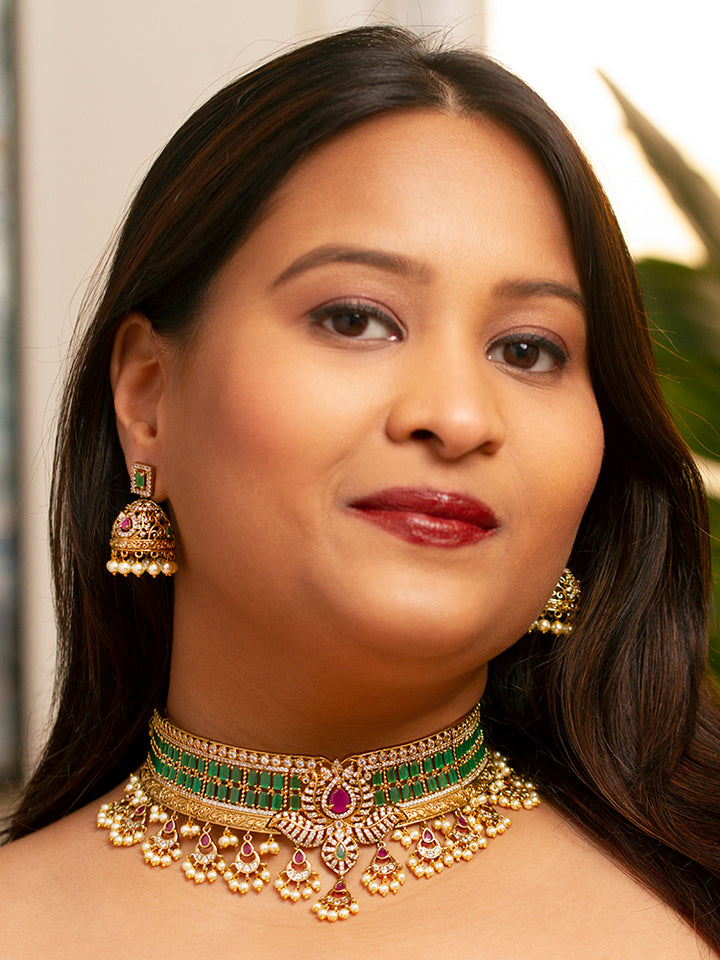 A close-up of a woman wearing the Kavira Choker Necklace Set—a green and pink necklace with gold details, green enamel, pink stones, pearl drops—paired with party earrings featuring pearls and a green gem.