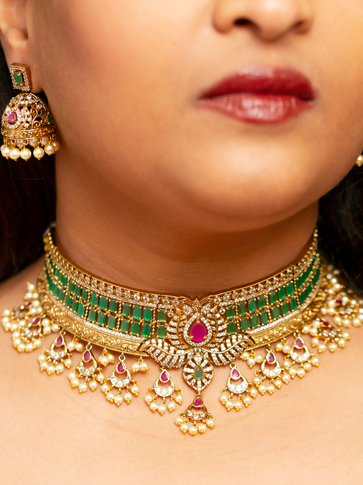 A close-up of a woman wearing the Kavira Choker Necklace Set—a green and pink necklace with gold details, green enamel, pink stones, pearl drops—paired with party earrings featuring pearls and a green gem.
