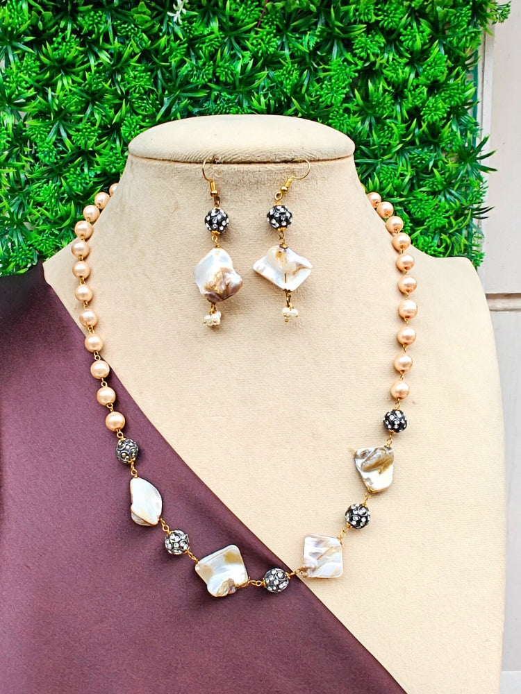 A beige bust displays the Kiara Necklace Set—gold-toned pearl necklace with matching dangling earrings featuring irregular white and gold beads, black rhinestone accents, and set against green artificial foliage.