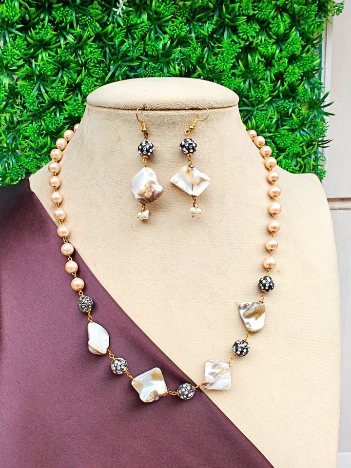 A beige bust displays the Kiara Necklace Set—gold-toned pearl necklace with matching dangling earrings featuring irregular white and gold beads, black rhinestone accents, and set against green artificial foliage.