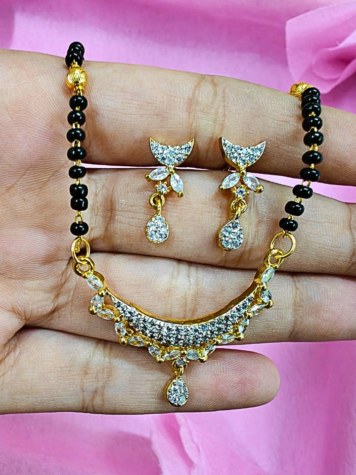 A hand holds the Kridha Mangalsutra—a gold necklace with black mangalsutra beads and matching earrings, pendant featuring sparkling American Diamonds—set against a soft pink background, perfect for your wedding ceremony dress. Diamond mangalsutra. Modern mangalsutra. 