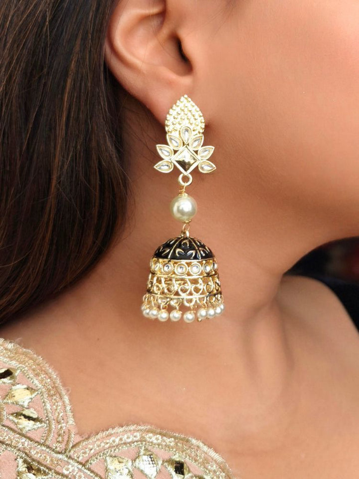 A close-up of a womans ear adorned with Kritika Black Earrings—large gold jhumkas featuring green meenakari work, pearl accents, kundan detailing, and a delicate floral moti near the earlobe.