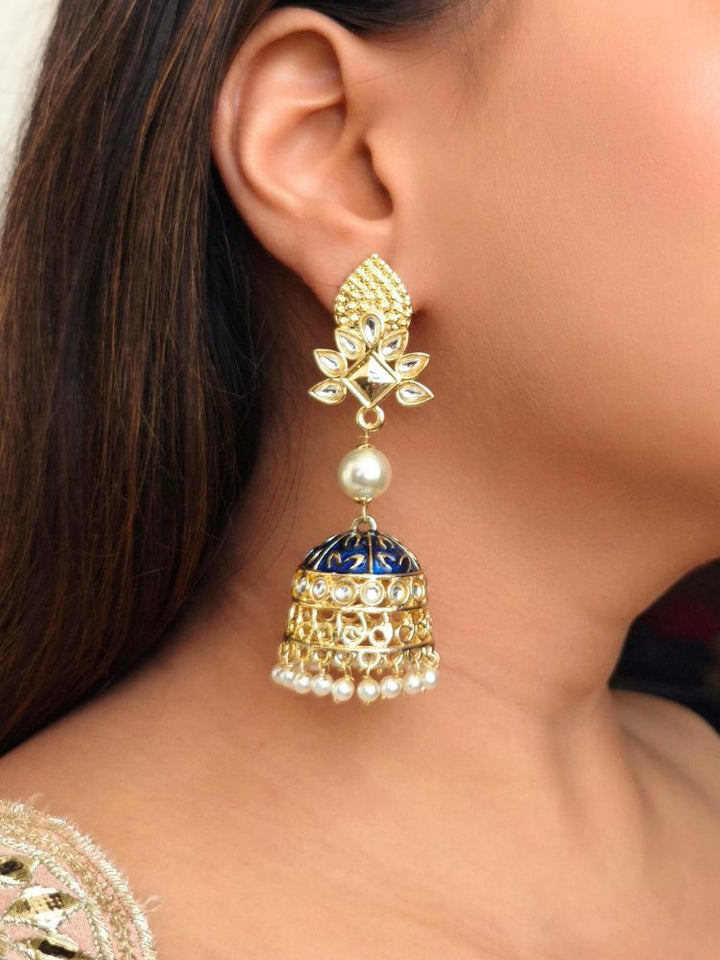 A close-up of a womans ear adorned with Kritika Blue Earrings—large gold jhumkas featuring green meenakari work, pearl accents, kundan detailing, and a delicate floral moti near the earlobe.