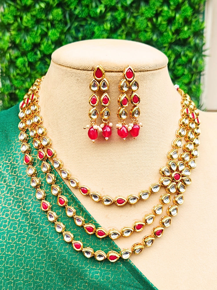 The Kumudini Red Necklace Set features three layers of red  gemstones and Kundan and matching dangling earrings, showcased on a mannequin in green fabric with a leafy backdrop. Ideal as a Kundan necklace wedding accessory.