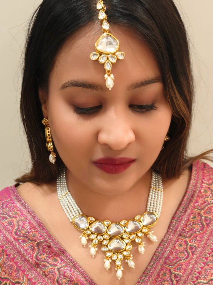 A woman in a pink embroidered saree wears the Kundan Vishakha Necklace Set, made with multiple stands of pears, big kundan stones, complemented by a maang tikka and matching earrings, closed eyes, neutral makeup, and bold red lipstick. Perfect shaadi necklace