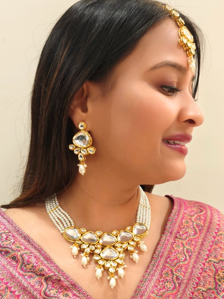 A woman in a pink embroidered saree wears the Kundan Vishakha Necklace Set, made with multiple stands of pears, big kundan stones, complemented by a maang tikka and matching earrings, closed eyes, neutral makeup, and bold red lipstick. Perfect shaadi necklace