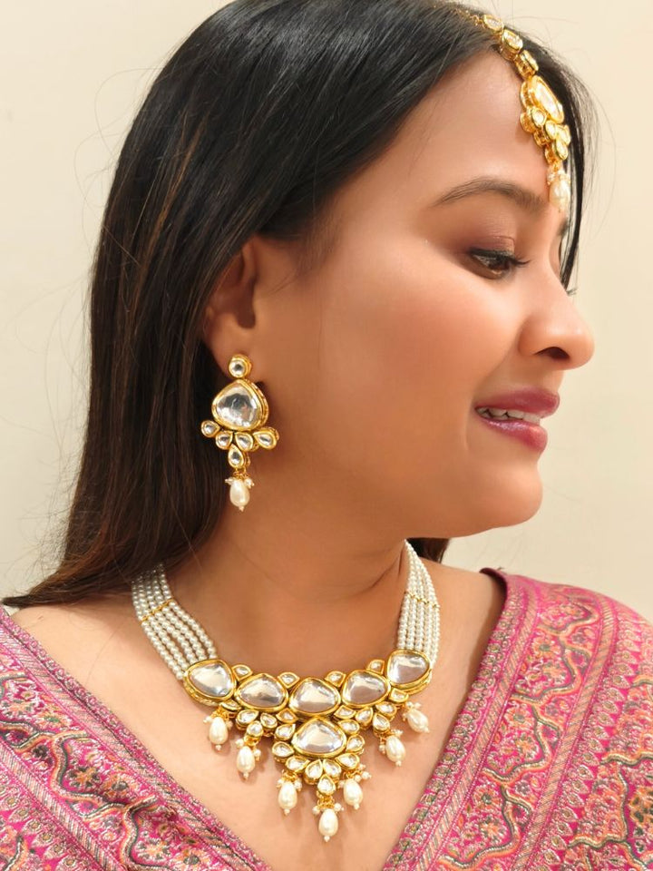 A woman in a pink embroidered saree wears the Kundan Vishakha Necklace Set, made with multiple stands of pears, big kundan stones, complemented by a maang tikka and matching earrings, closed eyes, neutral makeup, and bold red lipstick. Perfect shaadi necklace