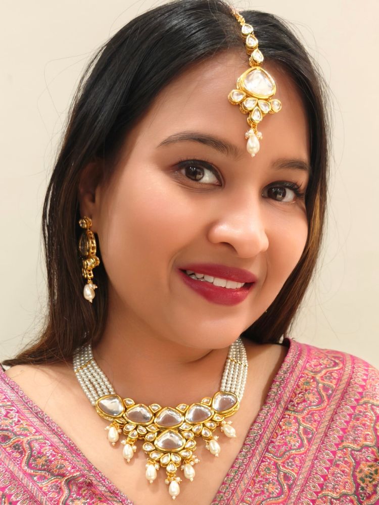 A woman in a pink embroidered saree wears the Kundan Vishakha Necklace Set, made with multiple stands of pears, big kundan stones, complemented by a maang tikka and matching earrings, closed eyes, neutral makeup, and bold red lipstick. Perfect shaadi necklace