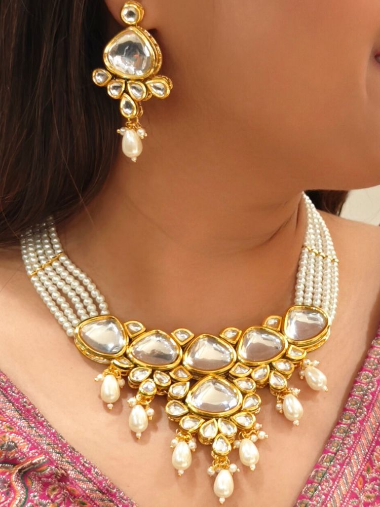 A woman in a pink embroidered saree wears the Kundan Vishakha Necklace Set, made with multiple stands of pears, big kundan stones, complemented by a maang tikka and matching earrings, closed eyes, neutral makeup, and bold red lipstick. Perfect shaadi necklace