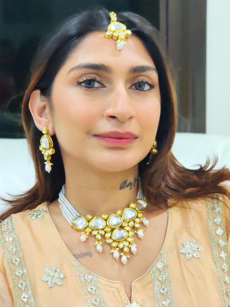 A woman in a Peach Outfit wears the Kundan Vishakha Necklace Set, made with multiple stands of pears, big kundan stones, complemented by a maang tikka and matching earrings, closed eyes, neutral makeup, and bold red lipstick. Perfect shaadi necklace