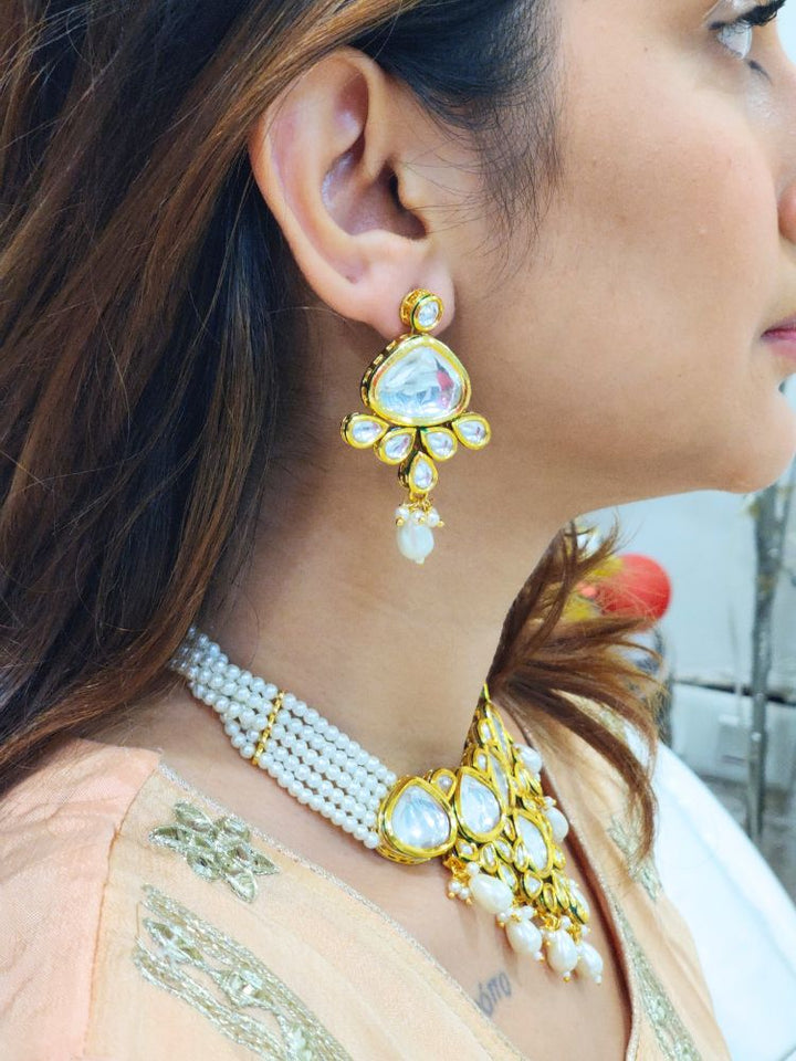 A woman in a Peach Outfit wears the Kundan Vishakha Necklace Set, made with multiple stands of pears, big kundan stones, complemented by a maang tikka and matching earrings, closed eyes, neutral makeup, and bold red lipstick. Perfect shaadi necklace