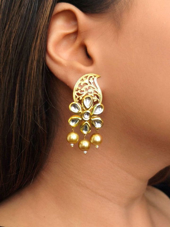 A womans ear adorned with the Kusum Earrings: large, ornate gold floral design with intricate filigree at the top, with Kundan stones shaped in a flower, along with three dangling gold pearls.