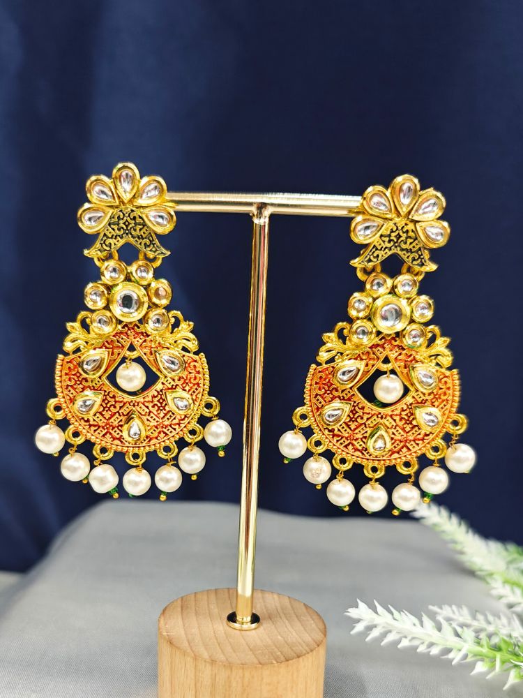 The Lajja Earrings are gold and red chandelier earrings featuring intricate Meenakari work, pearl accents, and teardrop stones, displayed on a stand against a dark blue background.