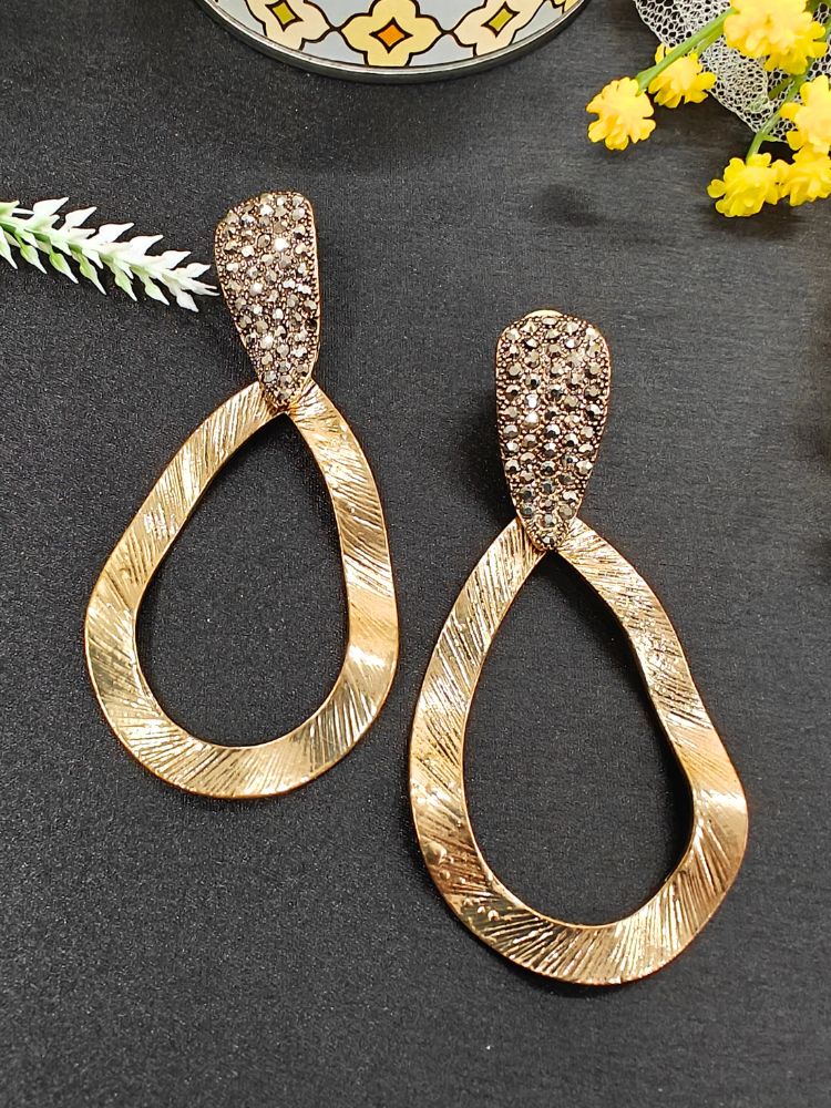 The Lara Earrings are gold-tone drop earrings featuring textured, wavy open oval shapes and crystal-studded tops—an elegant accessory to complement your wedding ceremony dress. Displayed on a black surface with decorative flowers.