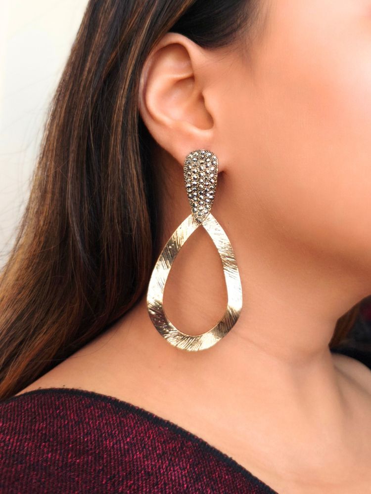 Close-up of a womans ear wearing  Lara Earrings are gold-tone drop earrings featuring textured, wavy open oval shapes and crystal-studded tops—an elegant accessory to complement your wedding ceremony dress.She has straight brown hair and is dressed in a dark maroon top.
