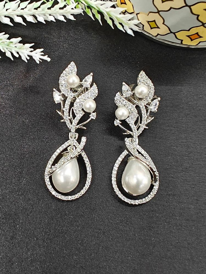 The Laurie Earrings, featuring ornate white leaf-shaped designs, pearl accents, and sparkling stones, capture the charm of western fashion as they rest on a black surface with decorative white foliage. Pearl Earrings, Date night jewellery. 