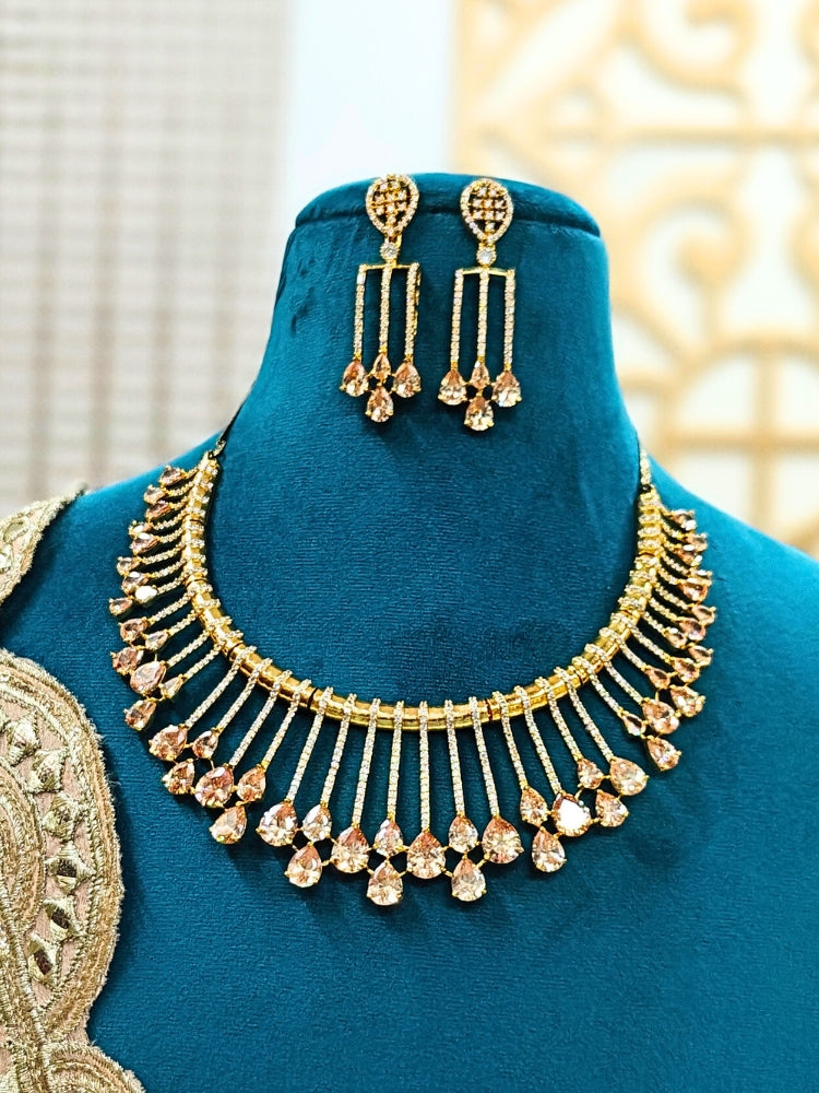 The Lavanya Necklace Set features a gold Champaign necklace with vertical bars topped by teardrop-shaped champagne colored American Diamonds, paired with matching dangling earrings and displayed on a blue velvet bust with ornate embroidery on the left.