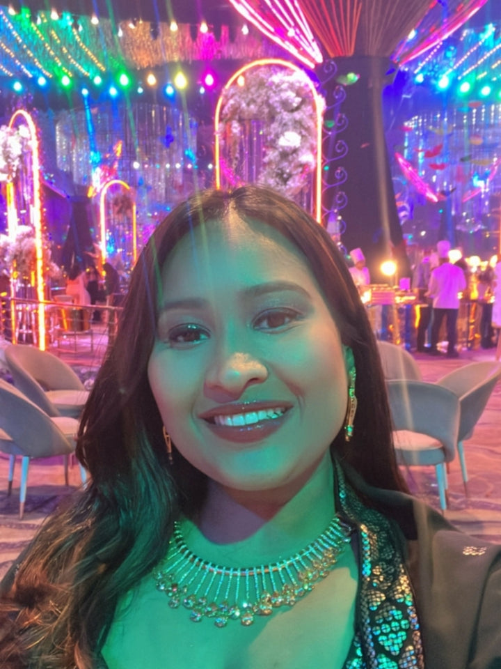 The Lavanya Necklace Set features a gold Champaign necklace with vertical bars topped by teardrop-shaped champagne colored American Diamonds, paired with matching dangling earrings and displayed on a blue velvet bust with ornate embroidery on the left.