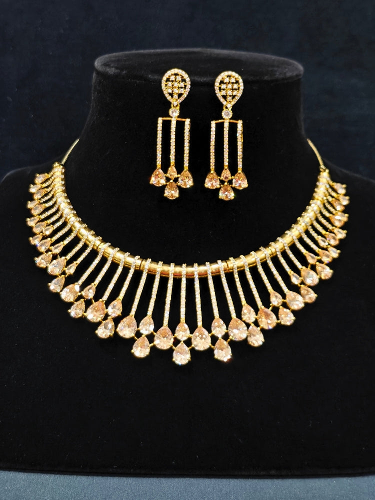 The Lavanya Necklace Set features a gold Champaign necklace with vertical bars topped by teardrop-shaped champagne colored American Diamonds, paired with matching dangling earrings and displayed on a blue velvet bust with ornate embroidery on the left.