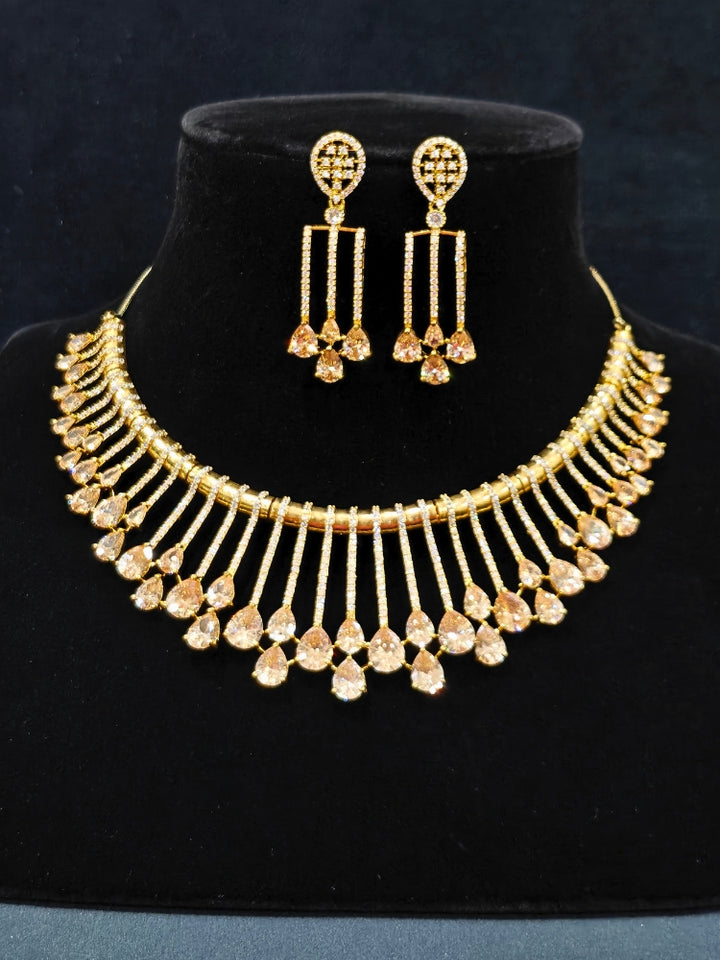 The Lavanya Necklace Set features a gold Champaign necklace with vertical bars topped by teardrop-shaped champagne colored American Diamonds, paired with matching dangling earrings and displayed on a blue velvet bust with ornate embroidery on the left.