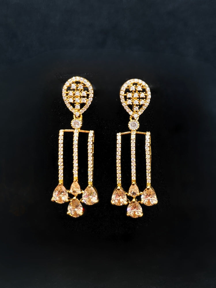 The Lavanya Necklace Set features a gold Champaign necklace with vertical bars topped by teardrop-shaped champagne colored American Diamonds, paired with matching dangling earrings and displayed on a blue velvet bust with ornate embroidery on the left.